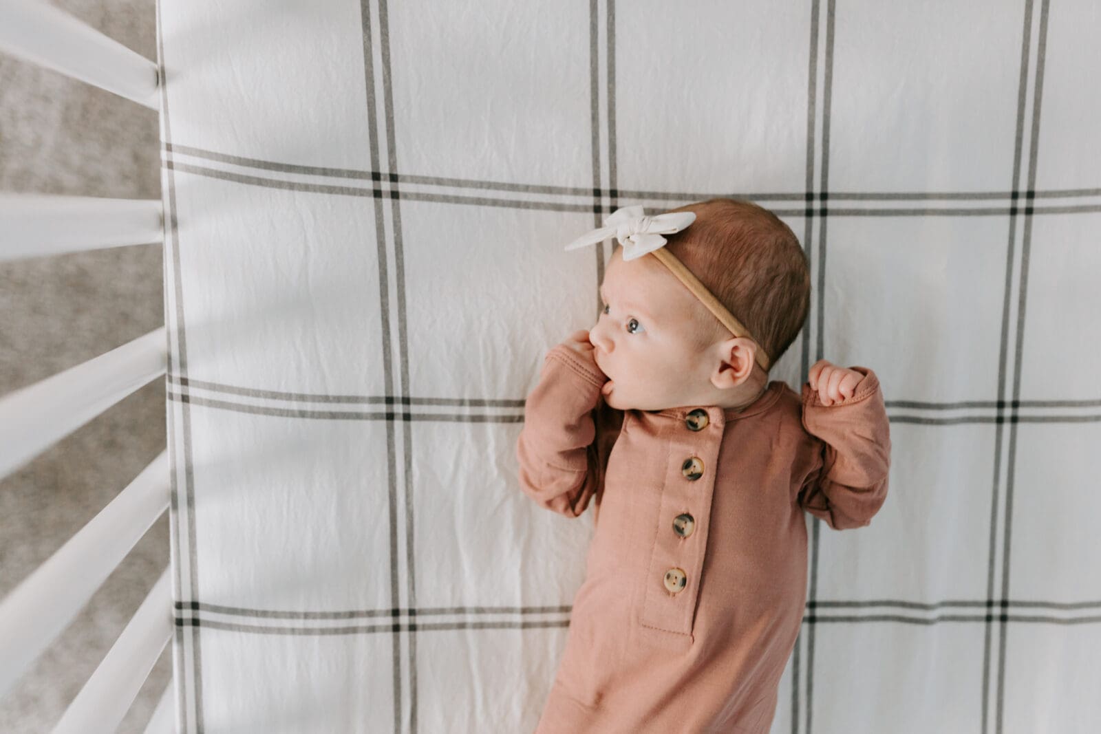 Newborn Photographer Vanessa Stoller captures an image from above of a newborn baby in a crib. The baby is in a pink sleeper, white bow, and is awake and looking to the side.