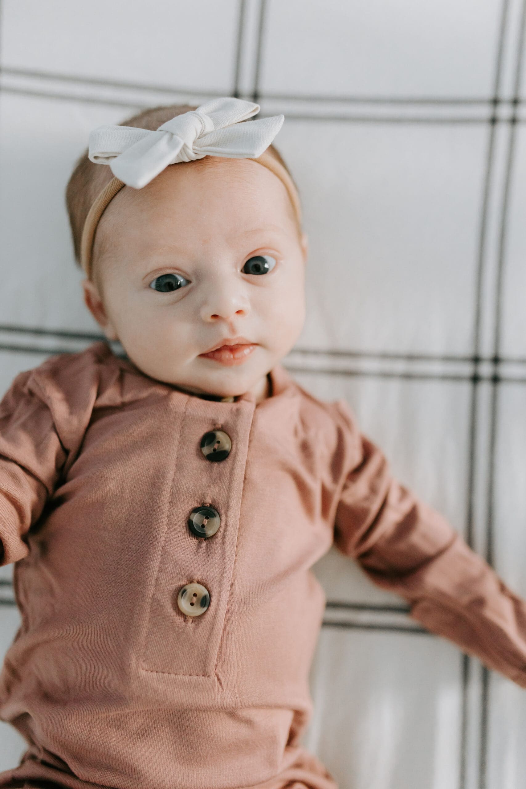 A newborn with bright eyes looks into the camera. She is wearing a pink sleeper with brown buttons and a white bow.