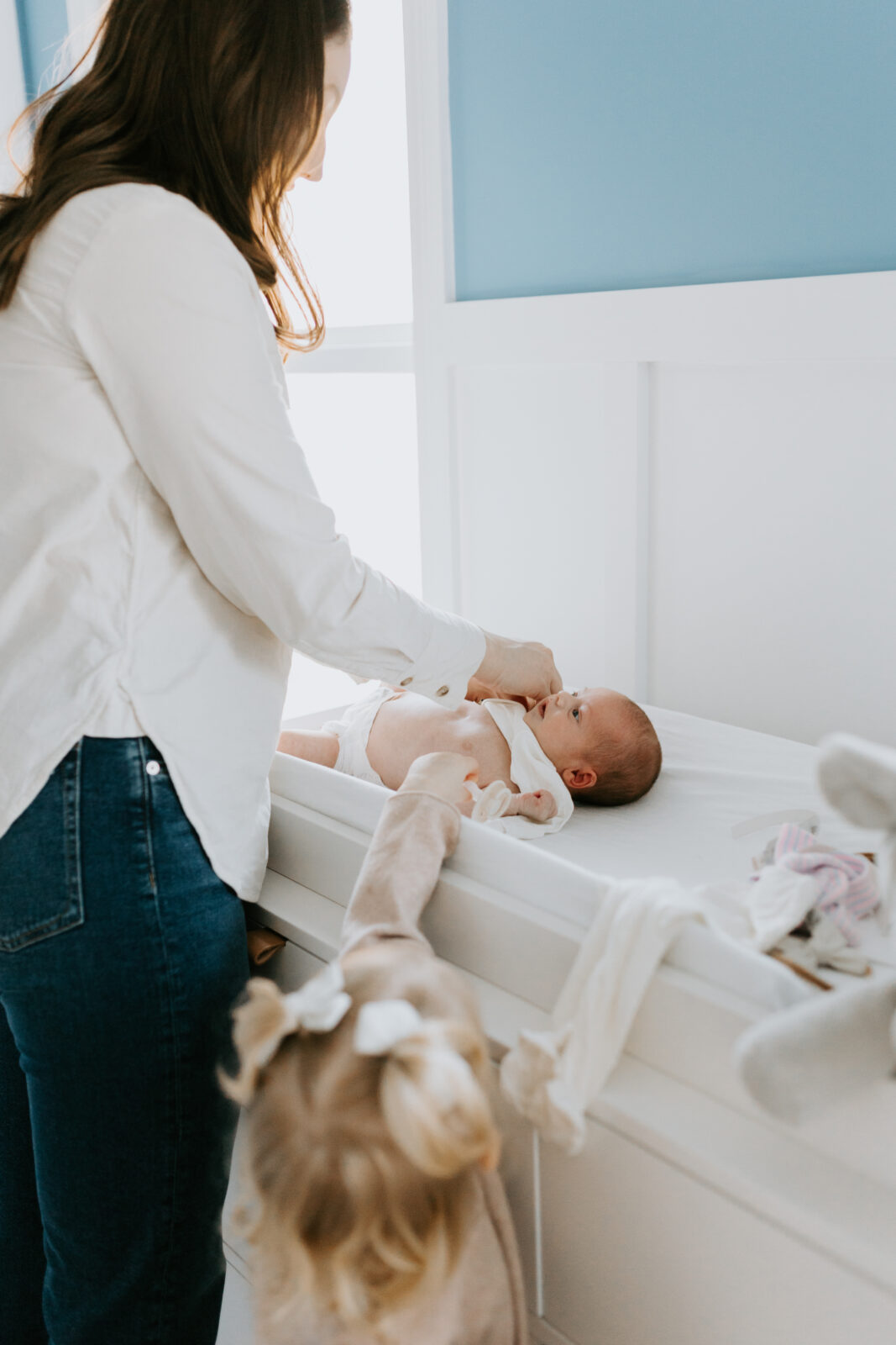 A mom stands at the changing table to change her newborn's outfit. A toddler stands nearby watching.