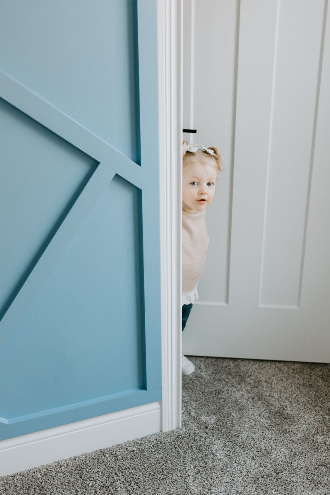 A toddler walks into the room of her baby sister's nursery. The room is blue and white. She stands at the threshold of the door looking in.
