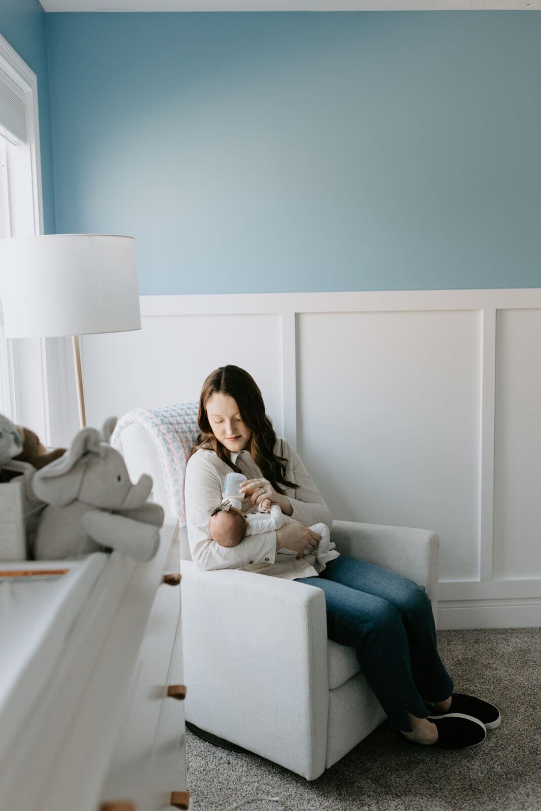 A mom sits in a rocking chair in her baby's blue nursery and feeds the baby a bottle. The window light falls on her face.