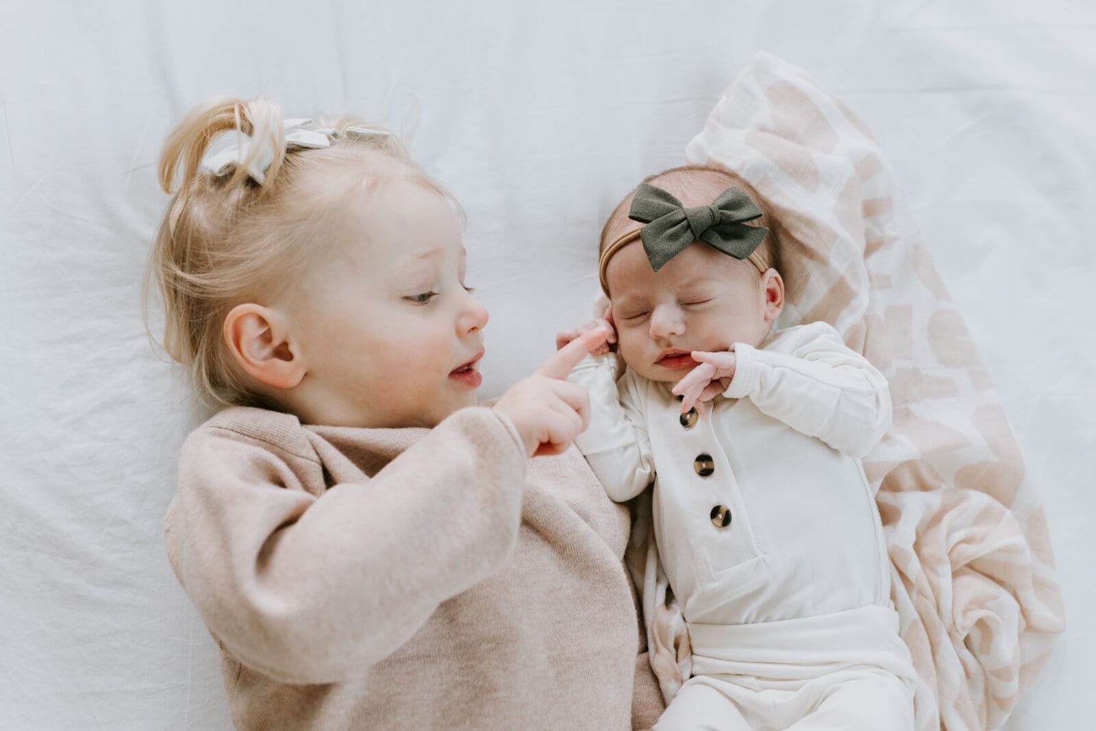A baby photo session with Vanessa Stoller Photography. This image captures a big sister laying next to and pointing at her baby sister.