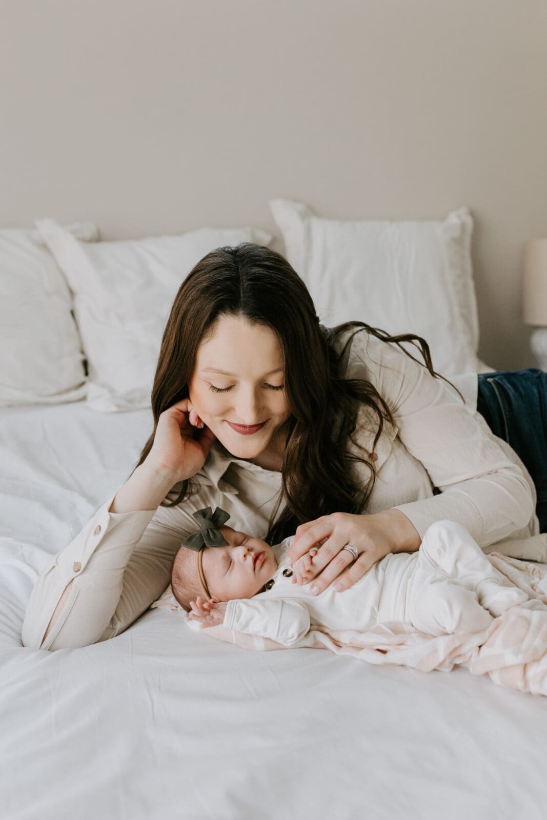 A Lafayette, Indiana motherhood photoshoot captures a mom laying on a white bed looking down at her sleeping newborn baby girl.