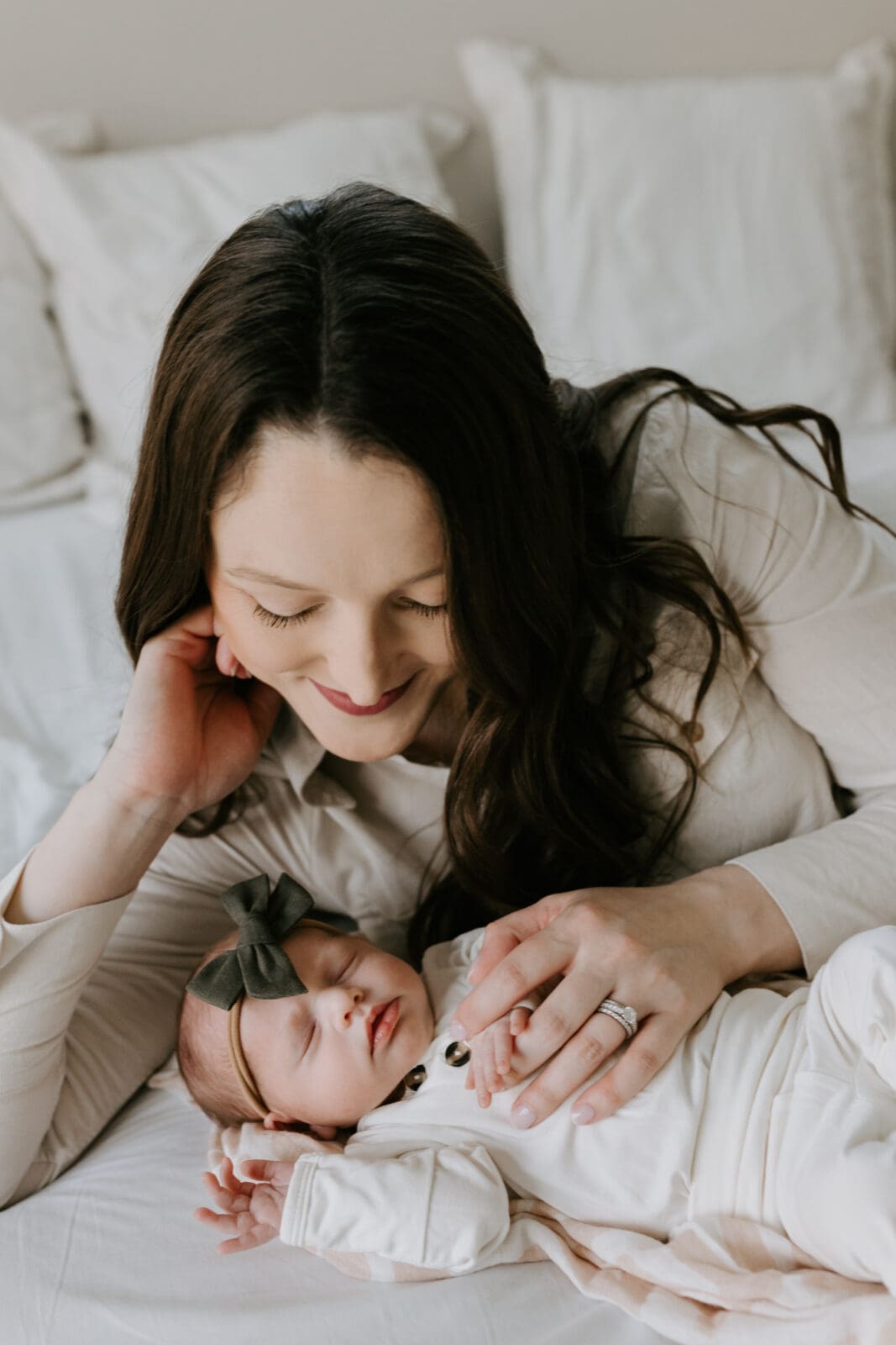 A mom with long brown gazes down and smiles at her newborn baby girl, gently placing her hand on the baby's chest.