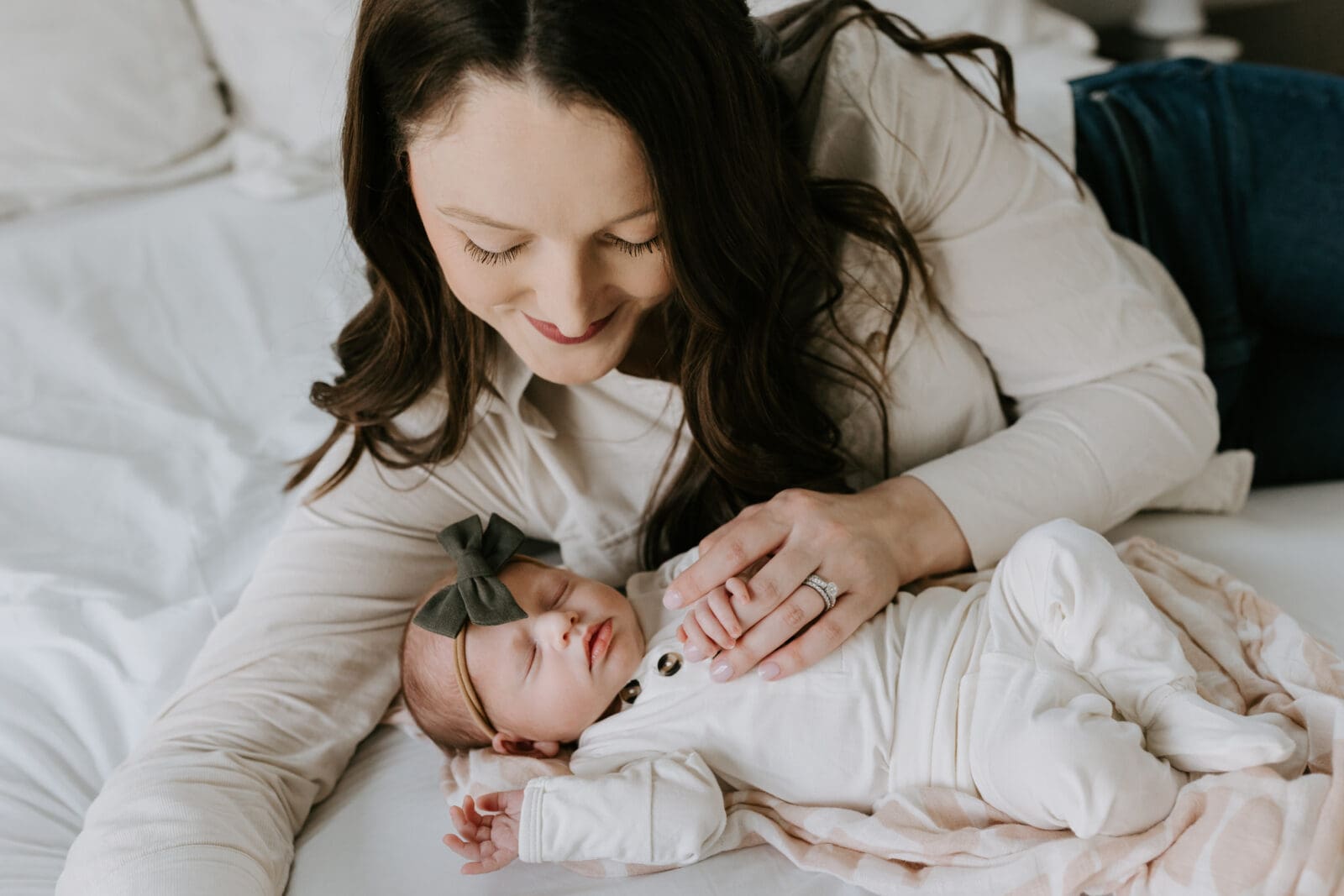 A mom lays near her infant baby girl and smiles down at her, holding the baby's tiny fingers in her hand. Newborn photography in Lafayette, Indiana.