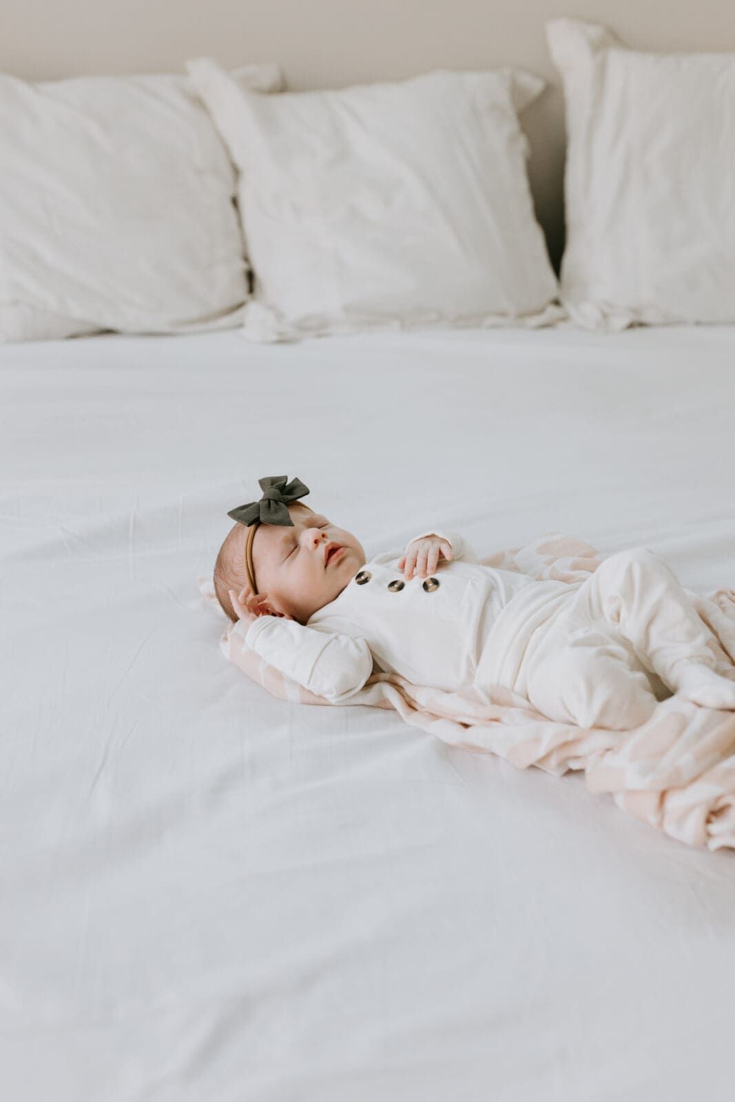A newborn baby girl wearing a white outfit and green bow lays on a white bed and sleeps.