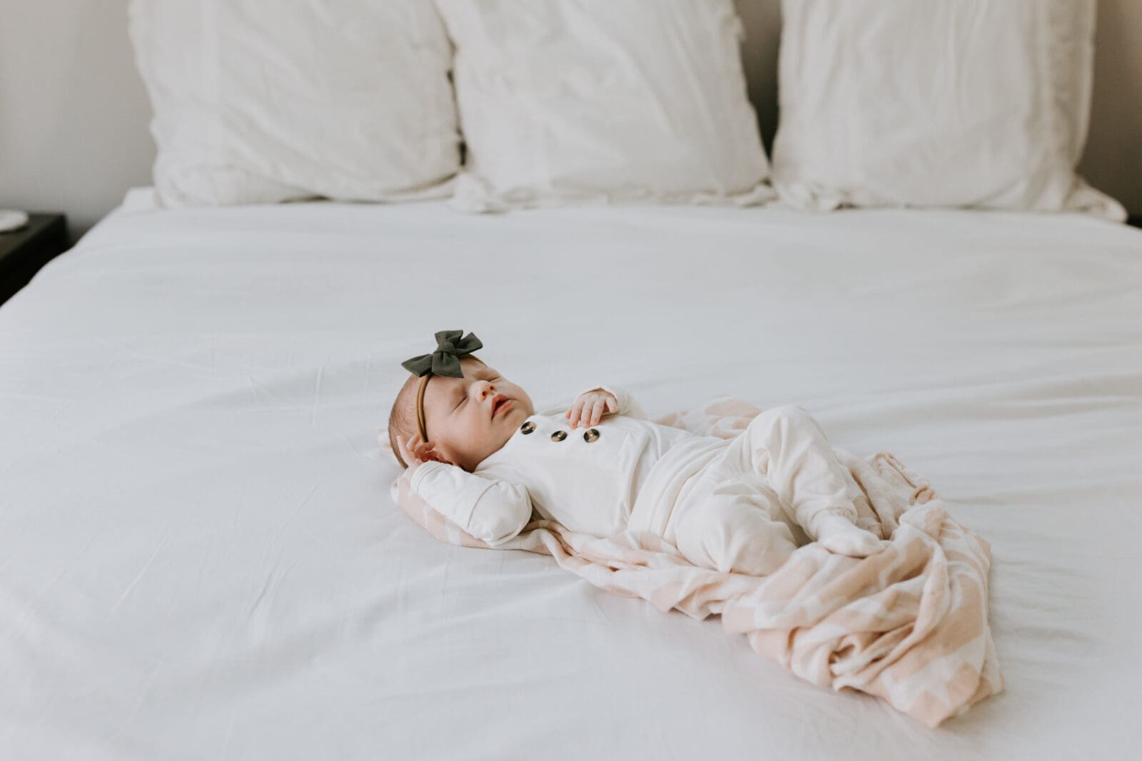 A newborn baby girl sleeps nestled in a swaddle blanket on a white bed. This image is lifestyle, as it captures the baby in her home, not in a studio.