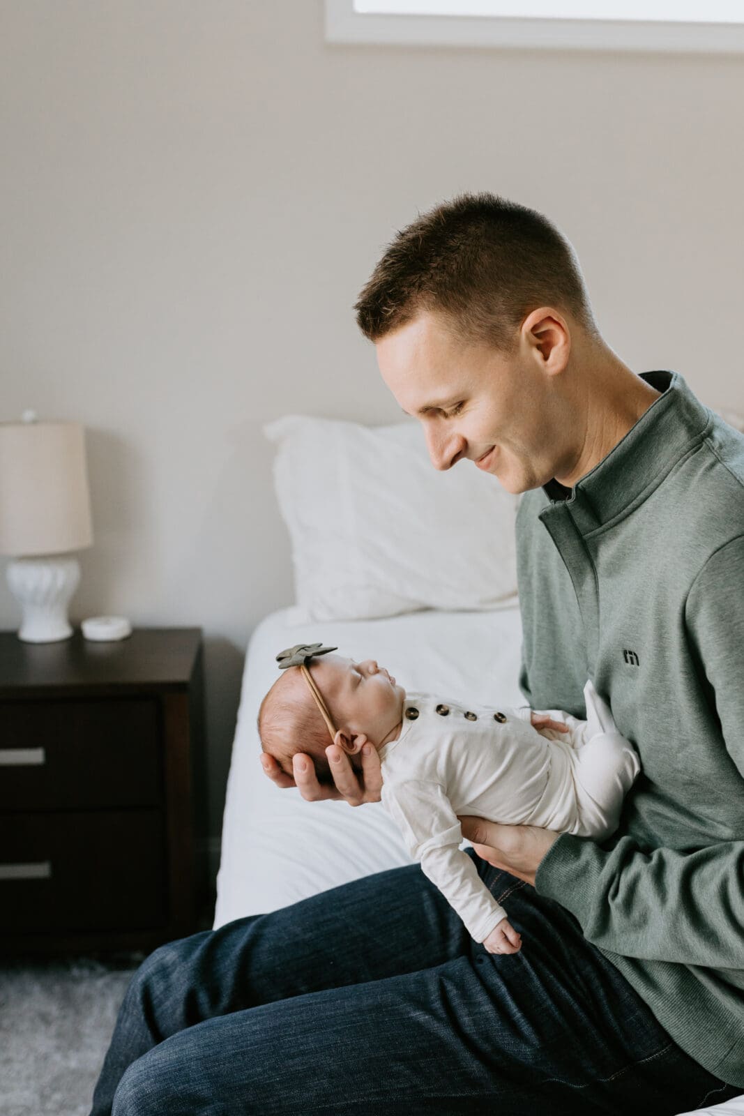 A dad sits on the edge of his white bed and smiles down at his newborn baby girl. He wears a green sweater and the baby wears a white outfit and green bow.