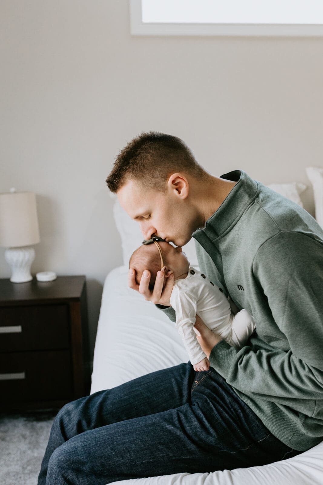 A dad sits on the edge of his white bed and kisses his baby girl on the head for this lifestyle newborn photography session with Vanessa Stoller Photography.