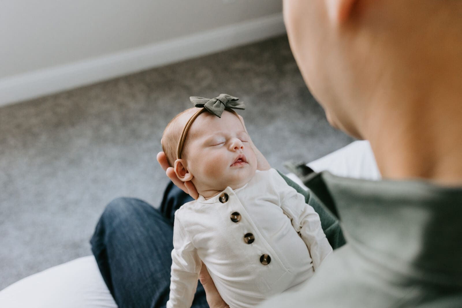 A dad sits on the edge of his bed and holds his baby girl in his hands. The photo angle is from over the shoulder of the dad looking down at the sleeping baby.