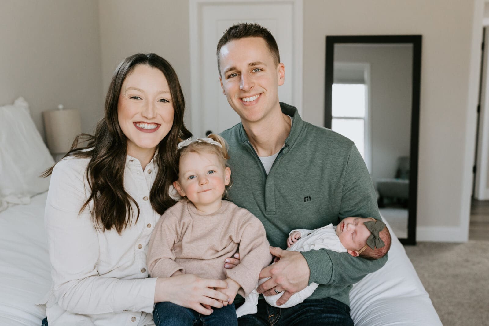 A lifestyle family photo of a family of four in their West Lafayette, Indiana bedroom. The mom and dad hold a toddler and newborn baby. All smile at the camera for Vanessa Stoller Photography.
