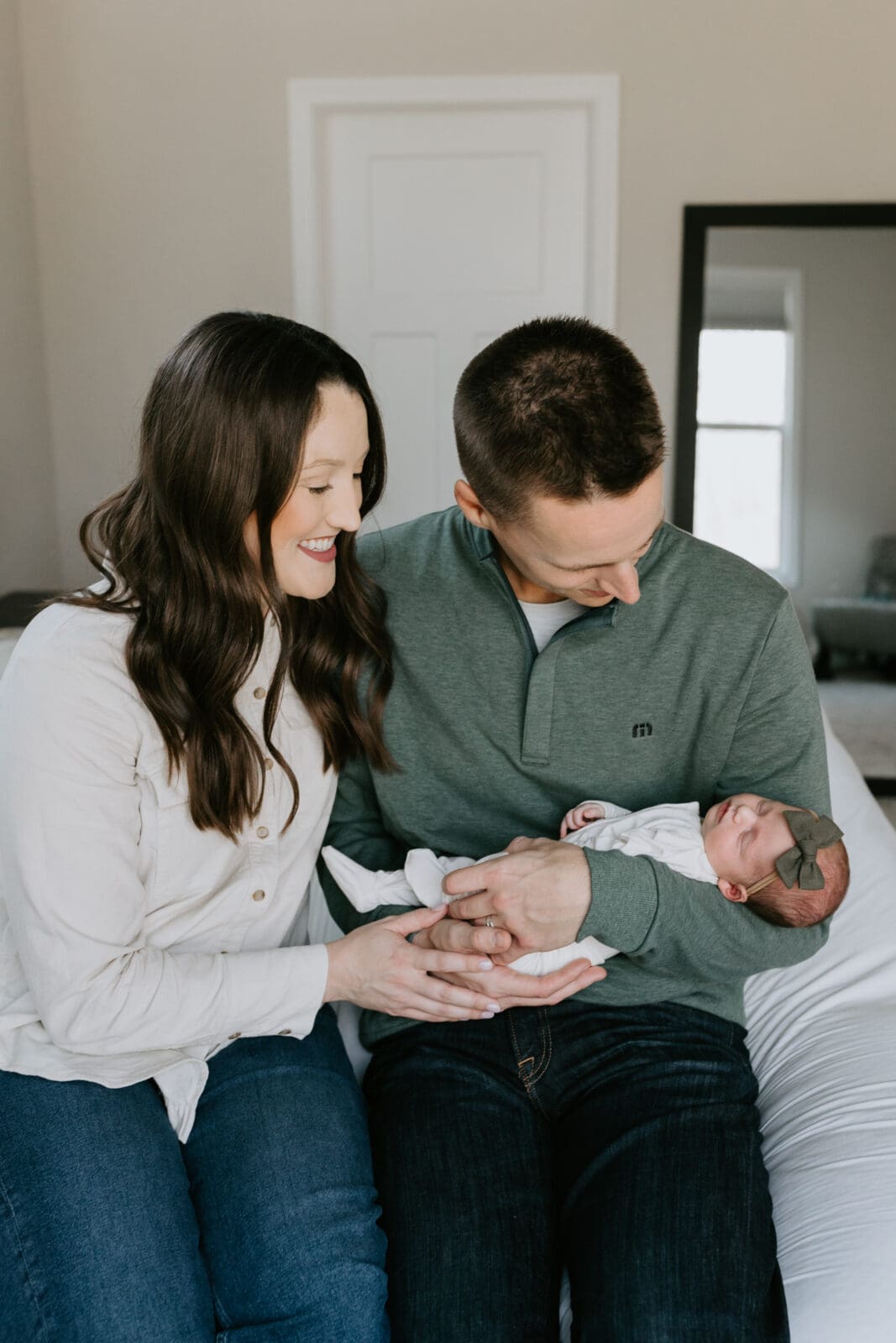 An in home newborn photo session in West Lafayette, Indiana captures a new dad and mom holding their newborn baby and lovingly gazing down at her. They sit on the edge of the bed in their bedroom.