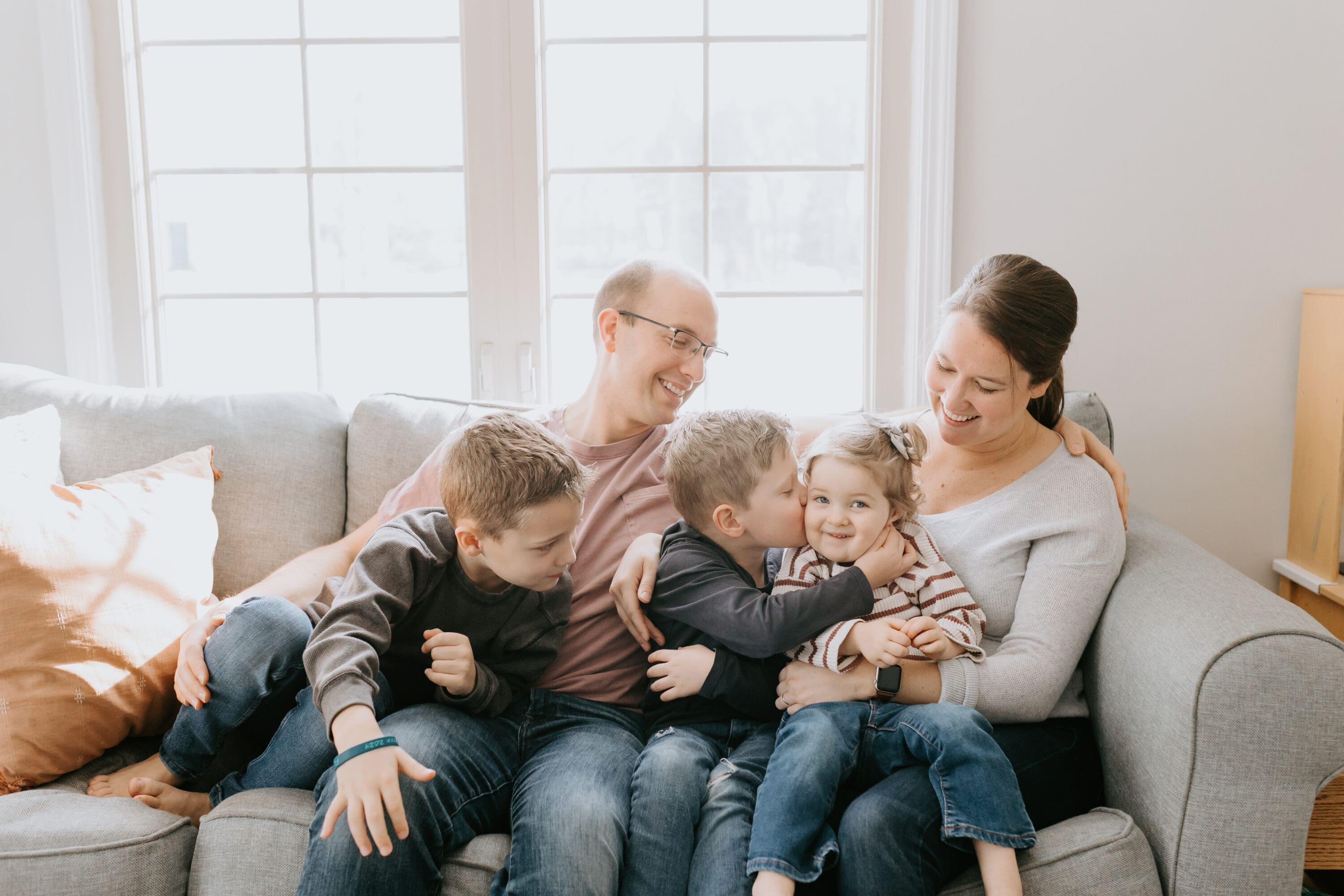A family cuddles on their sofa in front of a bright window. They look at each other and laugh. 